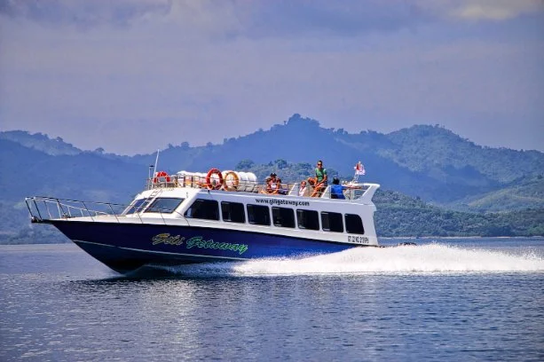 A white and blue bali fast boat sailing on a lake with mountains in the background, people onboard enjoying the ride.