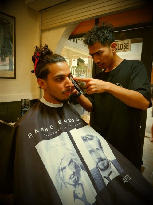 A man with curly hair and a beard gets a haircut at a barber shop while a barber cuts his hair using hair clippers. The man wears a cape with pictures of celebrities.
