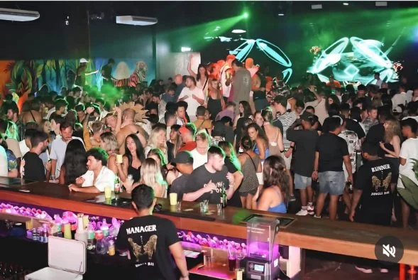Crowded nightclub with colorful lighting and large screen displaying abstract graphics, filled with people dancing and socializing.