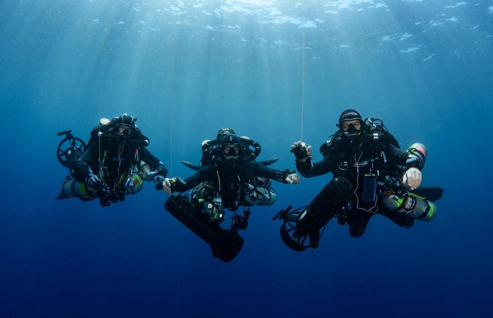 Three scuba divers underwater holding hands and waving at the camera.