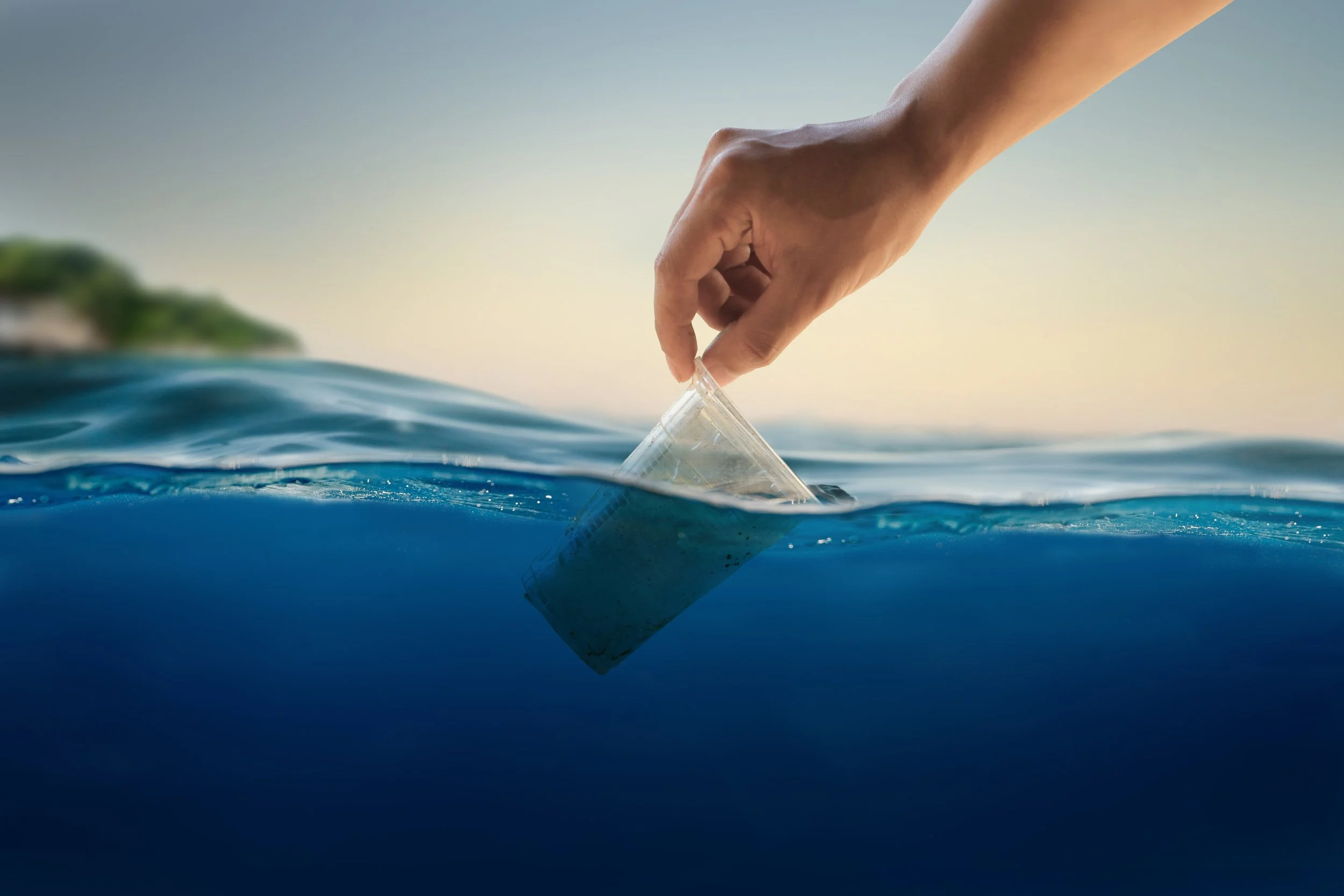 A hand is reaching into the ocean, holding a plastic cup, partially submerged in water with a blurred landmass and sky in the background.