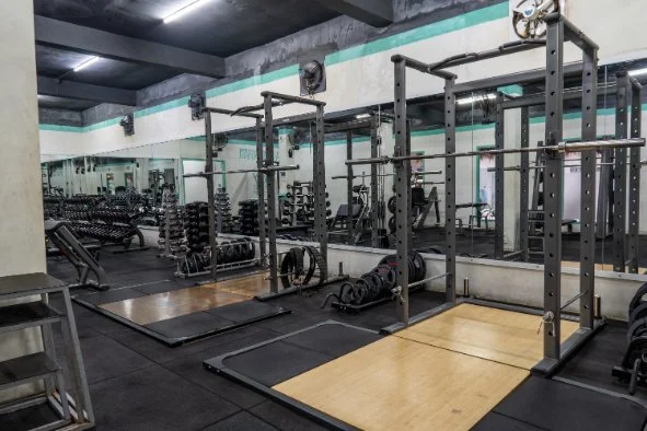 Empty gym with weightlifting equipment, including squat racks, weight plates, and mats, in a well-lit workout space.