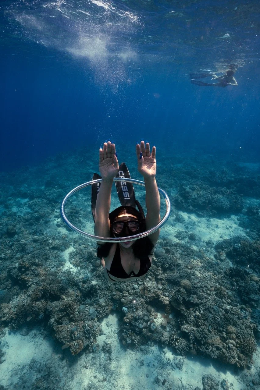 A woman wearing snorkeling gear, including a mask and a snorkel, swims underwater over a coral reef with her hands raised above her head, creating a circle with a hula hoop around her head. Another person is visible in the background, swimming near the surface of the water.