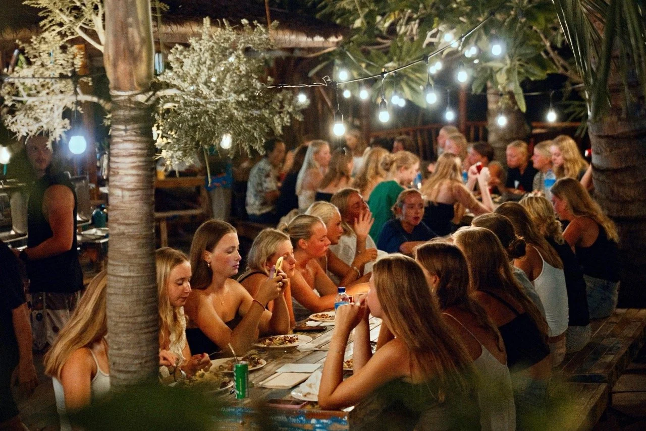 A group of young women sitting at a long outdoor wooden table under string lights at night, eating and socializing in a tropical setting with palm trees.