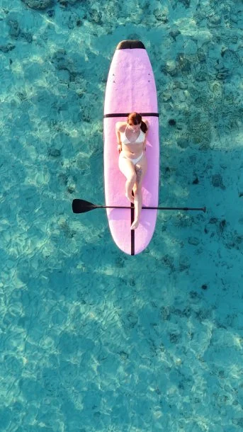 A woman in white swimsuit paddling a pink stand-up paddleboard on a clear blue ocean.