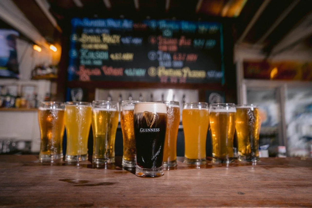 A row of ten beer glasses on a bar counter, with the center glass being a Guinness, set in a dimly lit pub with a chalkboard menu in the background.