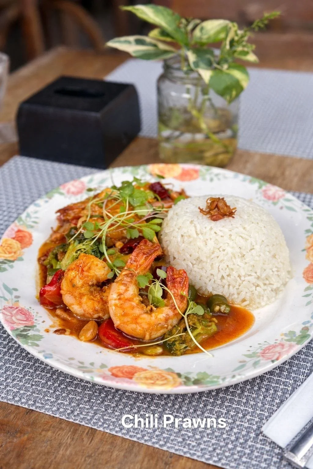 A plate of chili prawns with rice, garnished with microgreens, on a floral-patterned plate, set on a wooden table.
