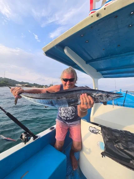 Person holding a large fish on a boat during daytime, with a view of the water and shoreline in the background.