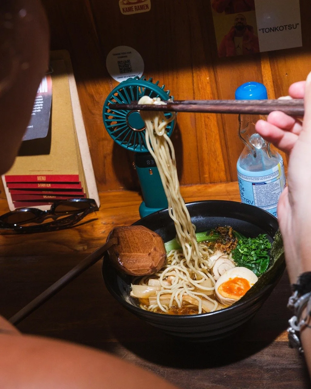 Person using chopsticks to lift ramen noodles from a bowl of ramen, which contains a soft-boiled egg, green onions, and broth, on a wooden table with a small portable fan, water bottle, sunglasses, and menu in the background.