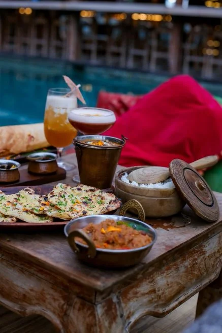 Outdoor table with Indian food and drinks near a swimming pool, including naan bread, a bowl of curry, cocktails, and a red cushion.