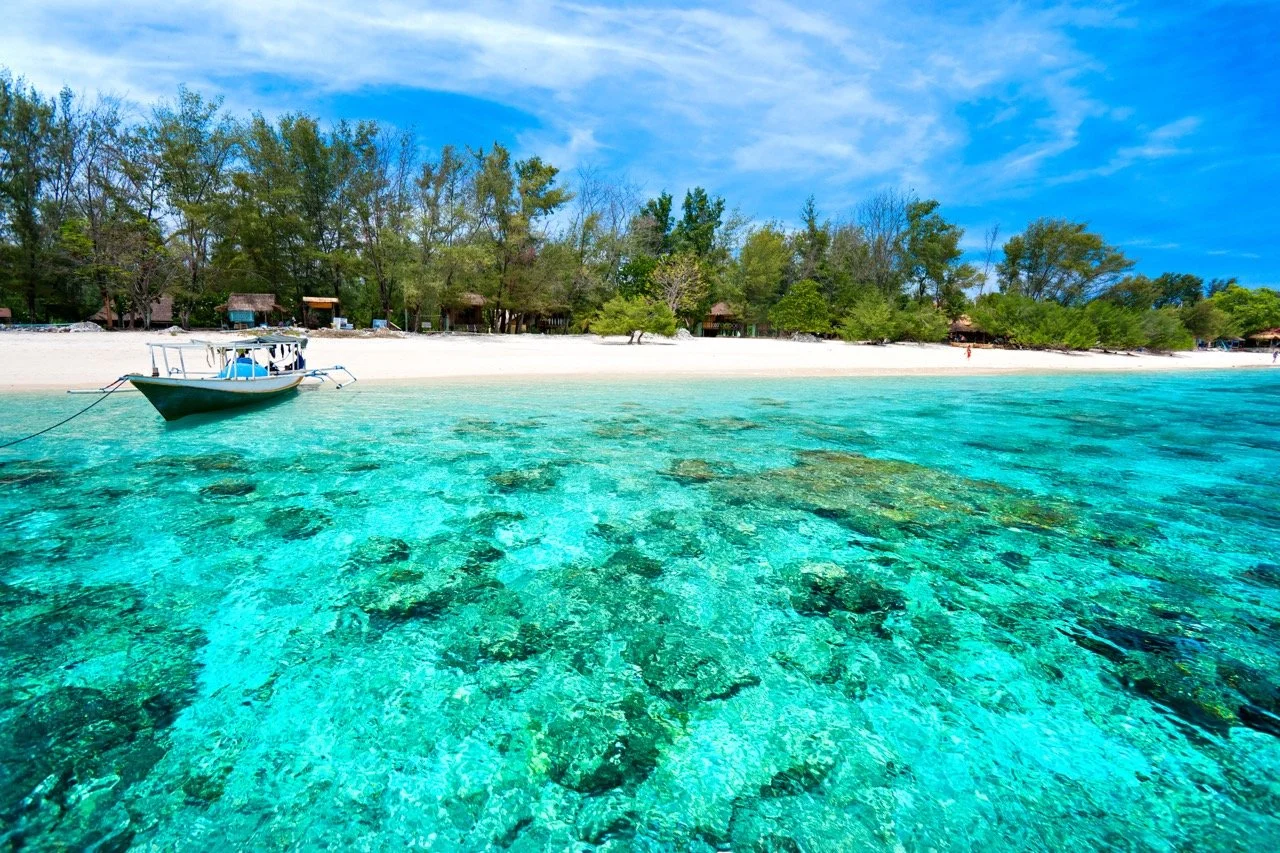 Tropical beach with white sand, clear turquoise water, and a small boat anchored near the shore. Trees and huts can be seen in the background under a partly cloudy sky.