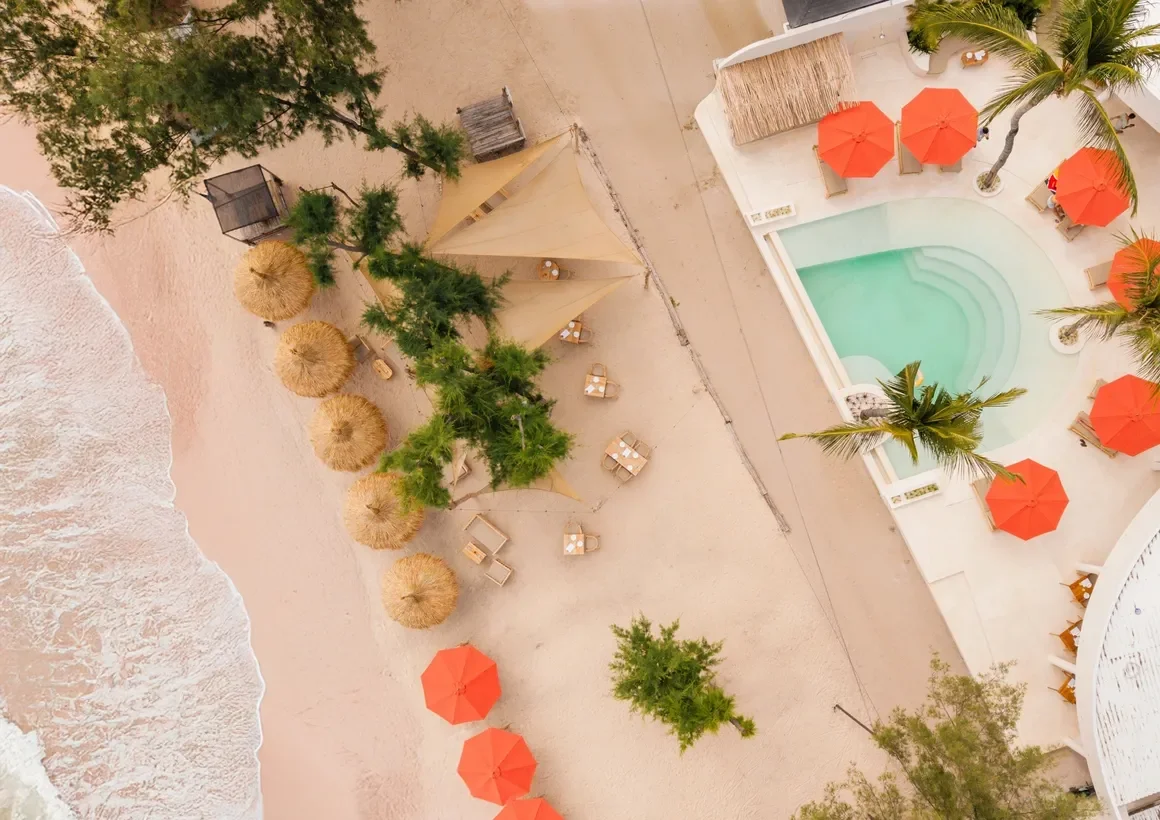 An aerial view of a beach resort area showing pink sand, a small swimming pool with surrounding orange umbrellas, and shaded seating areas with straw and umbrella shades, along with some trees.