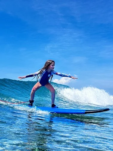 Young girl surfing on a blue surfboard in the ocean with clear blue sky above.
