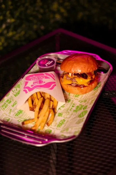 A tray with a cheeseburger, fries, and a small container of dipping sauce on a black metal table outdoors at night.