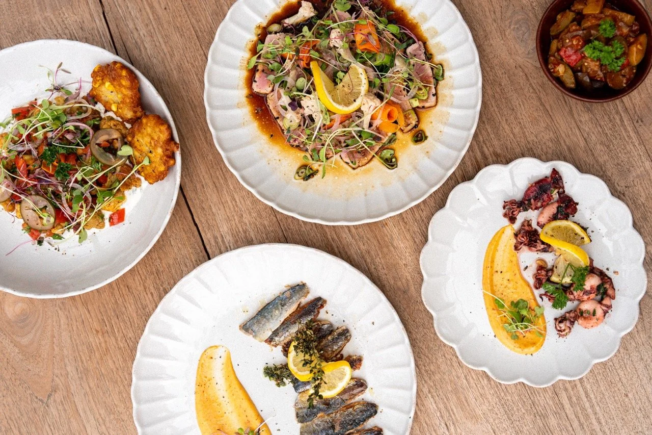 Assorted dishes on white plates and bowls, including a fish dish, a beef salad, a shrimp plate with lemon, a vegetable stir-fry, and a bowl of eggplant stew, all on a wooden table.