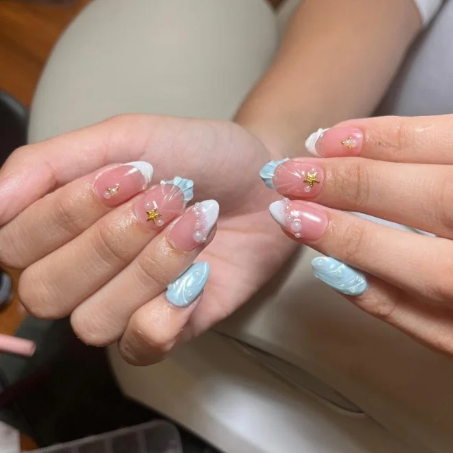 Close-up of hands showcasing decorated nails with pastel blue and white nail polish, gold star and crescent moon decals, and pearl accents.