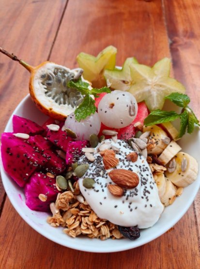Bowl of sliced dragon fruit, star fruit, dragon fruit, banana, granola, and yogurt decorated with seeds, nuts, and fresh mint leaves placed on a wooden table.