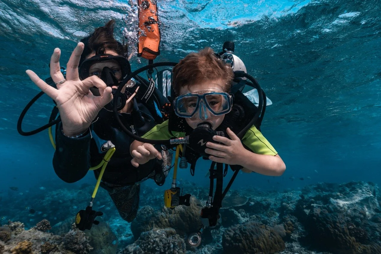 Two scuba divers underwater, one showing an okay sign and the other holding a mask to their face.