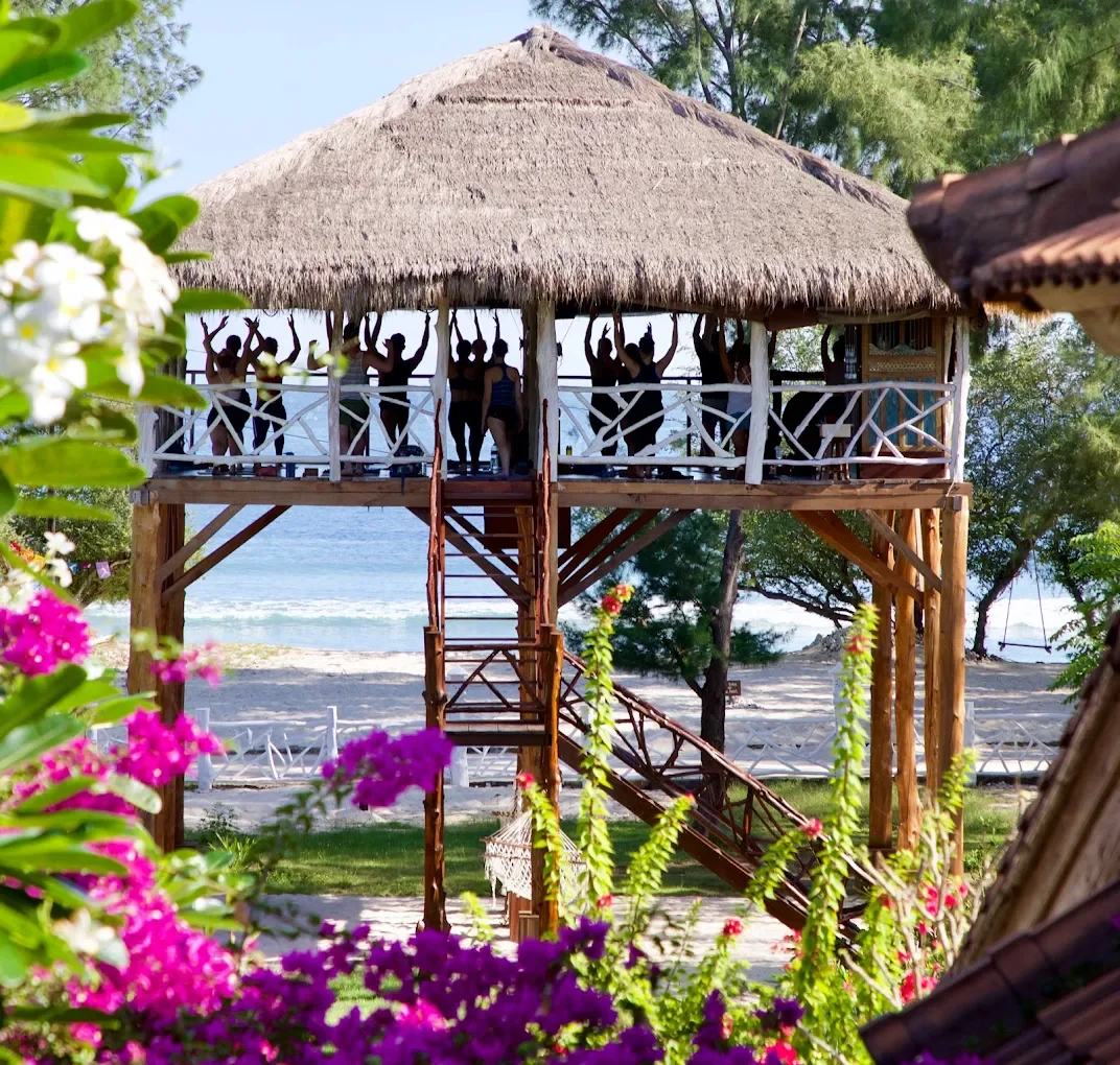 Group of people practicing yoga on a balcony of a thatched-roof hut at the beach, surrounded by trees and colorful flowers.