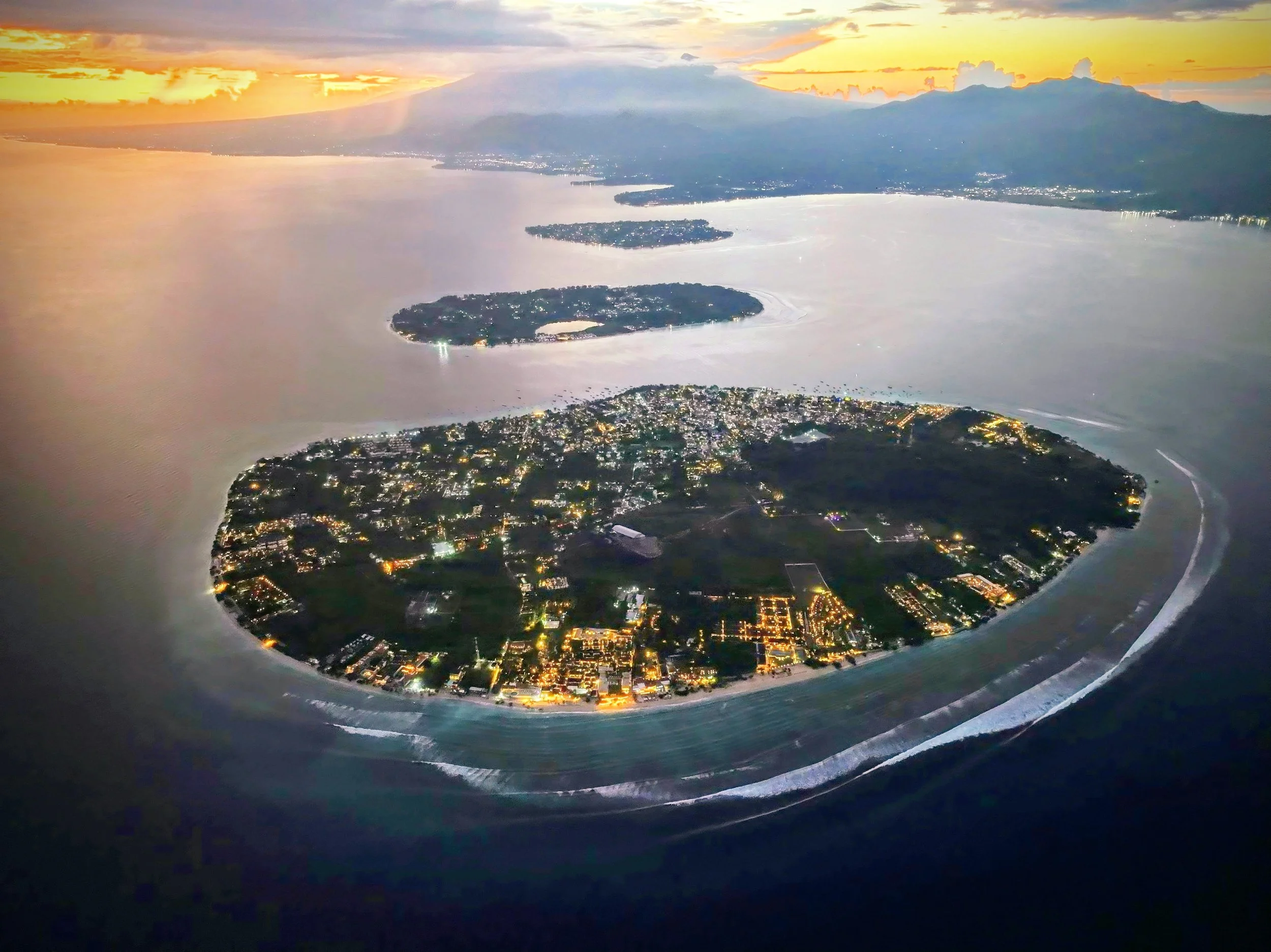 Aerial view of an island with a city at sunset, surrounded by water and a volcanic mountain in the background.