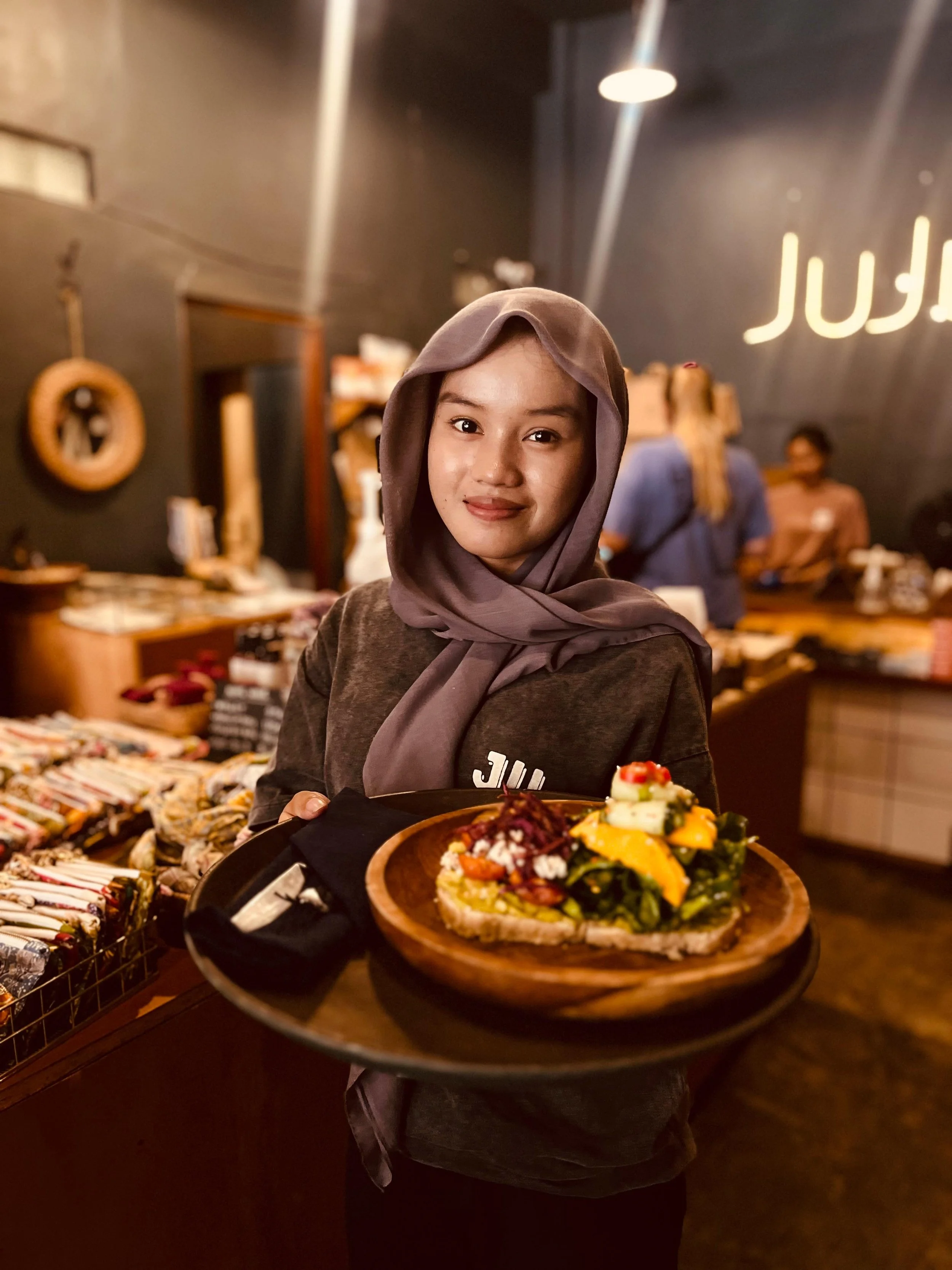 A woman with a gray headscarf holding a plate of avocado toast topped with vegetables and cheese in a food store.