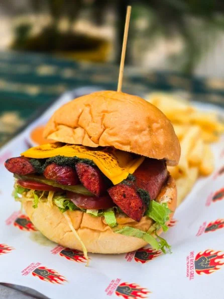 Close-up of a sandwich with sausage, cheddar cheese, lettuce, and a bun, served on a paper-lined tray with fries in the background.