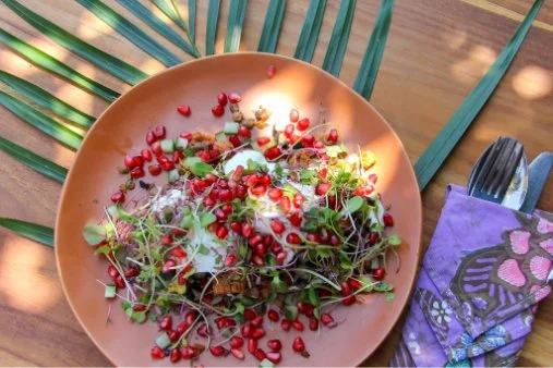 A coral-colored plate filled with pomegranate seeds and microgreens, with palm leaves, a fork, and a purple napkin nearby.