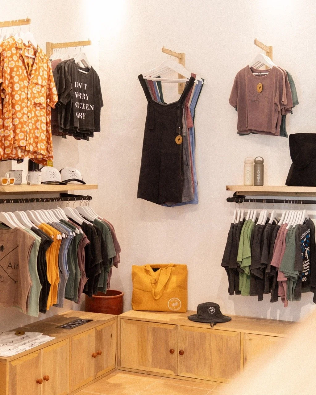 Clothing store display with t-shirts, tank tops, and accessories on wooden shelves and hangers, including a yellow tote bag and a black bucket hat.