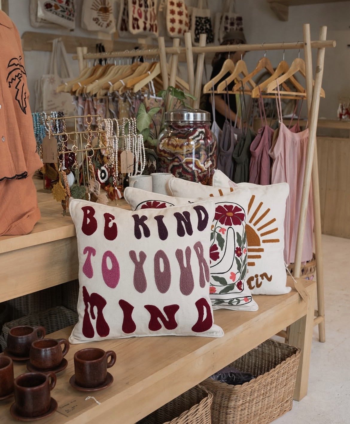 Decorative pillows on a wooden shelf, one with the phrase 'Be Kind to Your Mind' in colorful letters, another with a floral design and sun motif, surrounded by jewelry, ceramics, and clothing items in a boutique store.