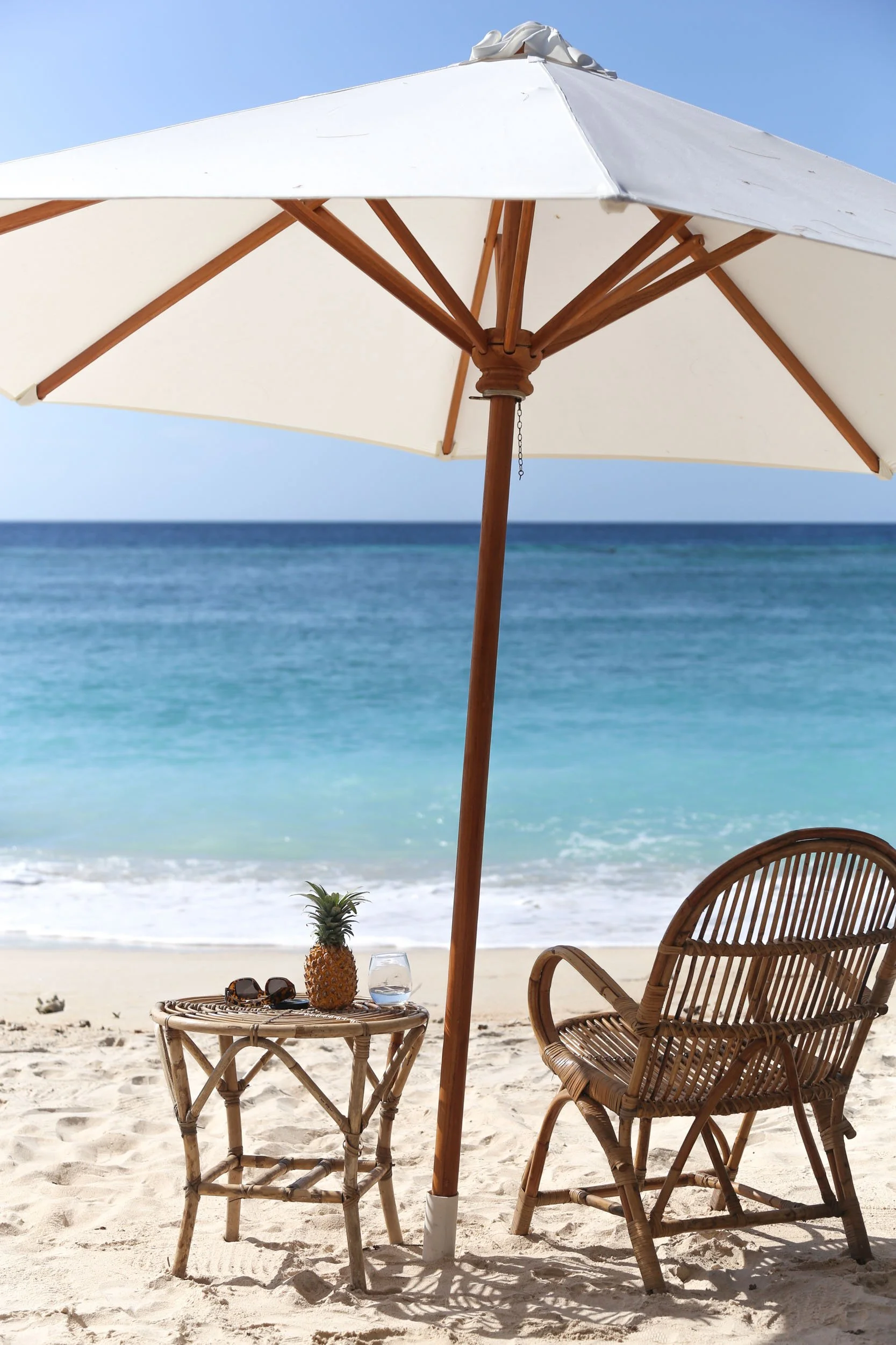 A beach scene with a white umbrella, two wicker chairs, a small wicker table with sunglasses, a pineapple, a glass of water, and a scenic ocean in the background.