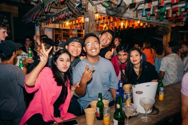Group of young people enjoying a night out at a bar or party, with some making peace signs and smiling, surrounded by drinks and a lively crowd.