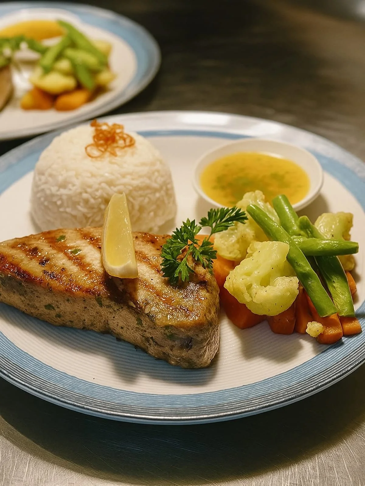 A plate of grilled fish with lemon wedge, white rice, mixed vegetables, and a small bowl of yellow soup on a decorative blue and white plate.