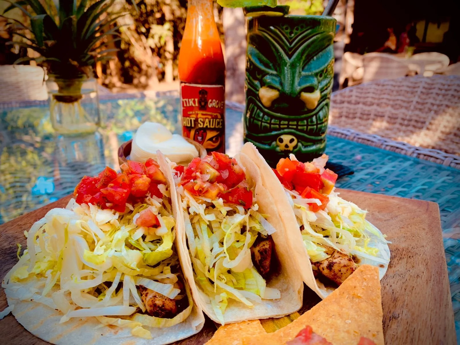Three tacos filled with lettuce, grilled meat, and salsa, served on a wooden board, with a bottle of hot sauce, a wedge of onion, and a decorative tiki mug in the background on a blue table outdoors.