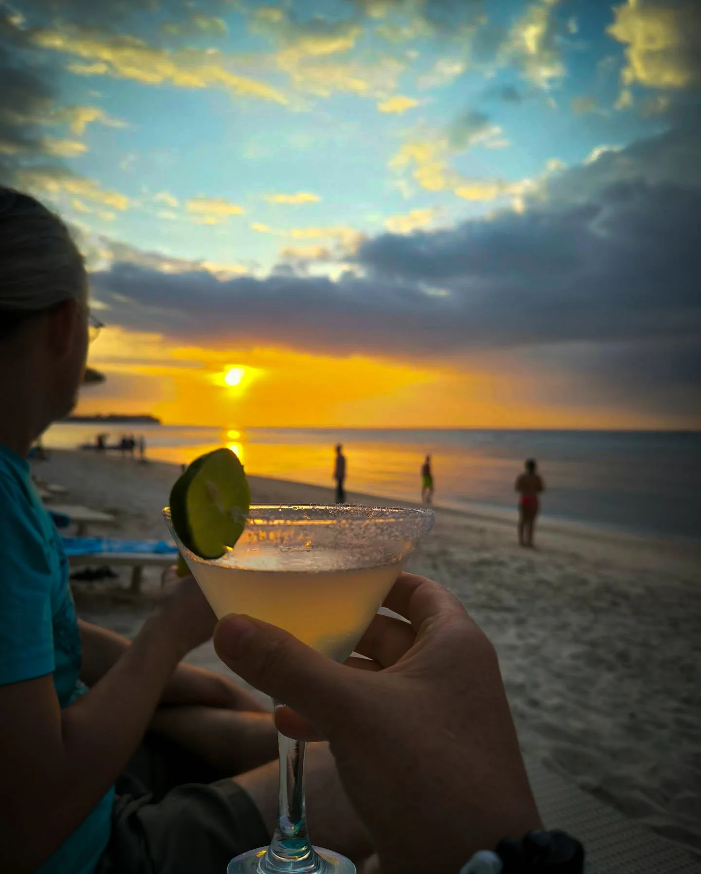 Person holding a cocktail glass with lime at a beach during sunset, with several people in the background near the shoreline.