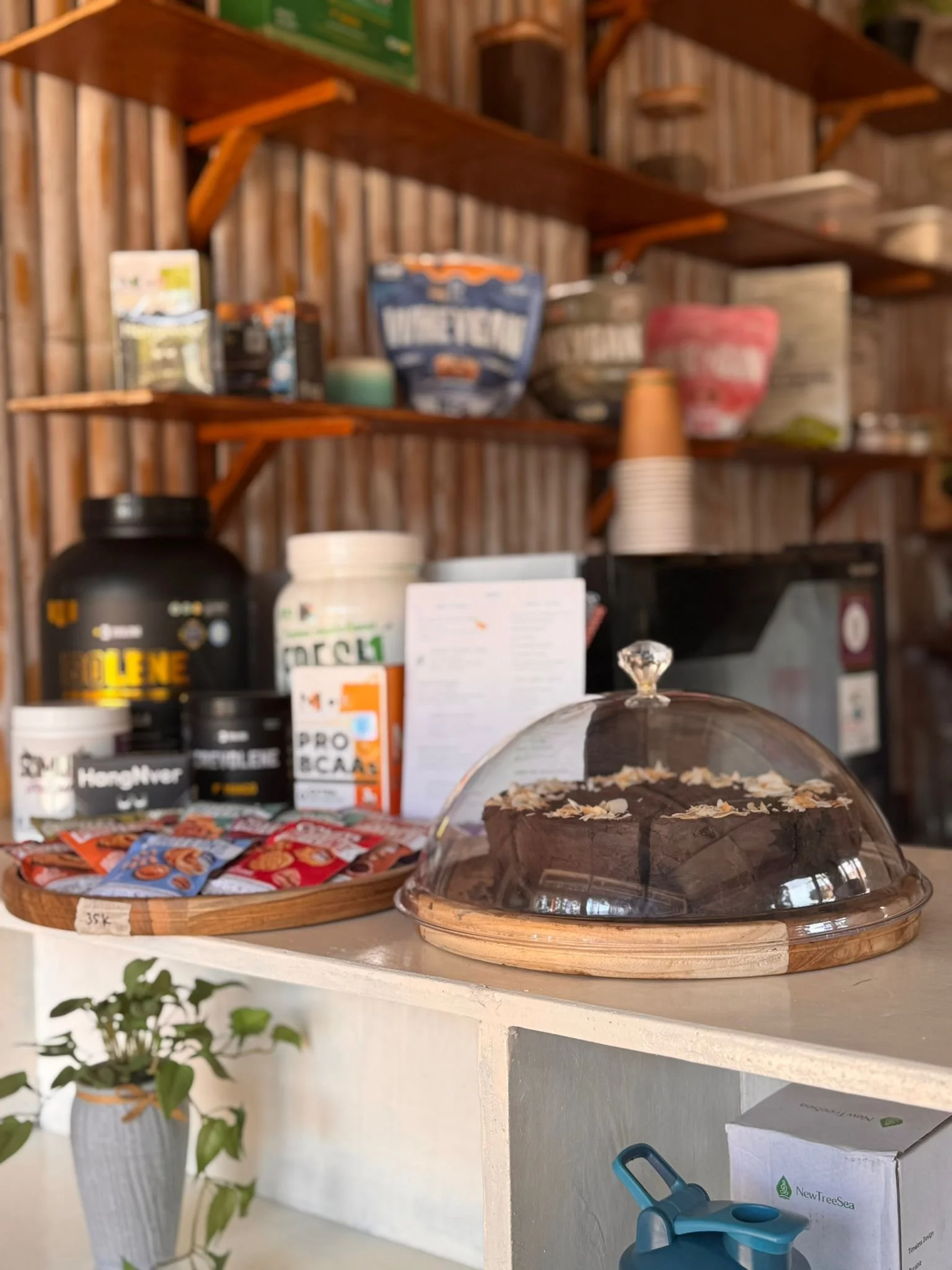 A cake with chocolate frosting and almond slices inside a glass dome on a wooden tray, placed on a white counter with assorted protein supplements and snack packets behind it.