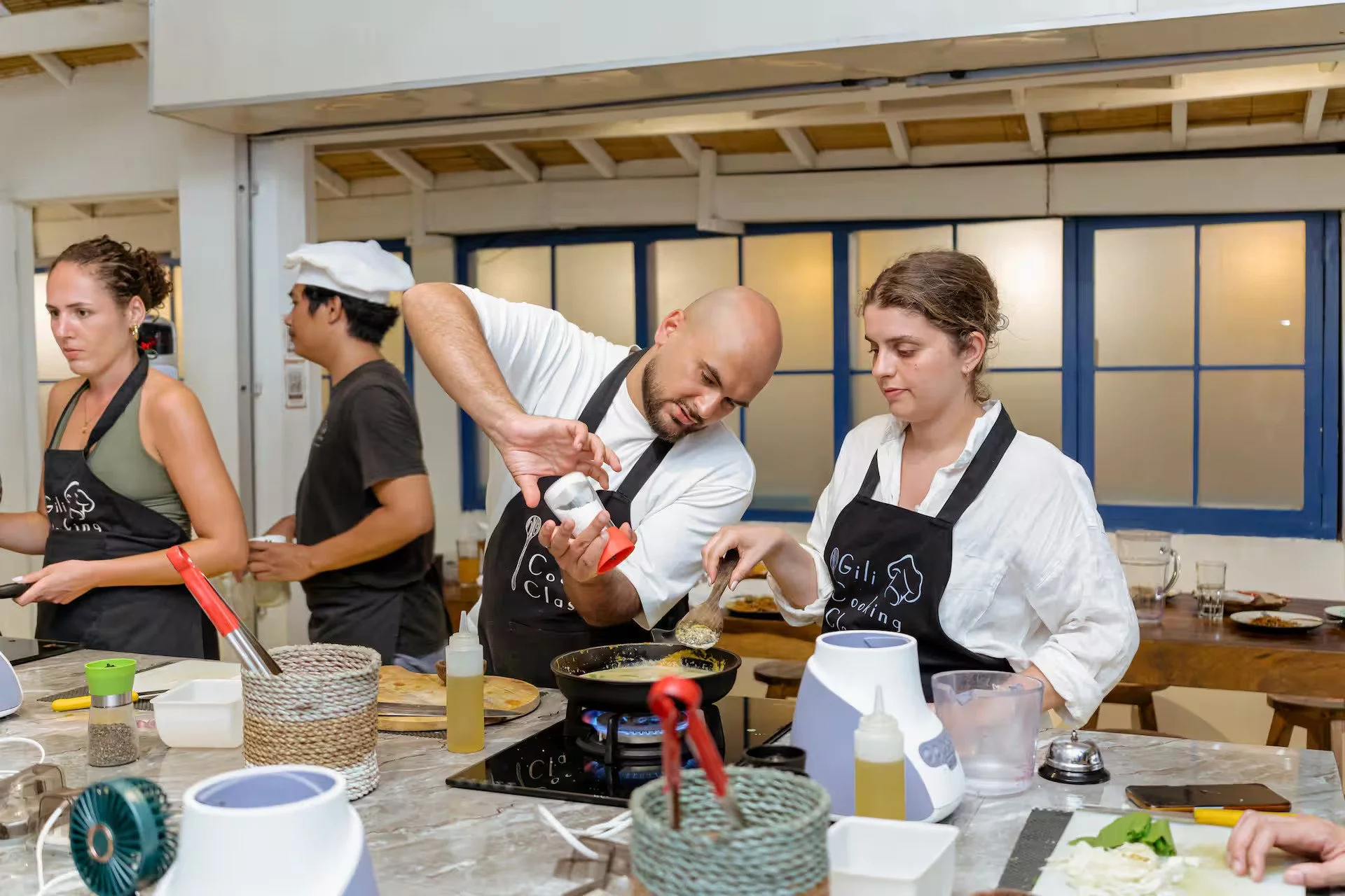 People at Gili cooking classes, in a kitchen, with one person pouring seasoning into a pan while another observes, surrounded by cooking utensils and ingredients.