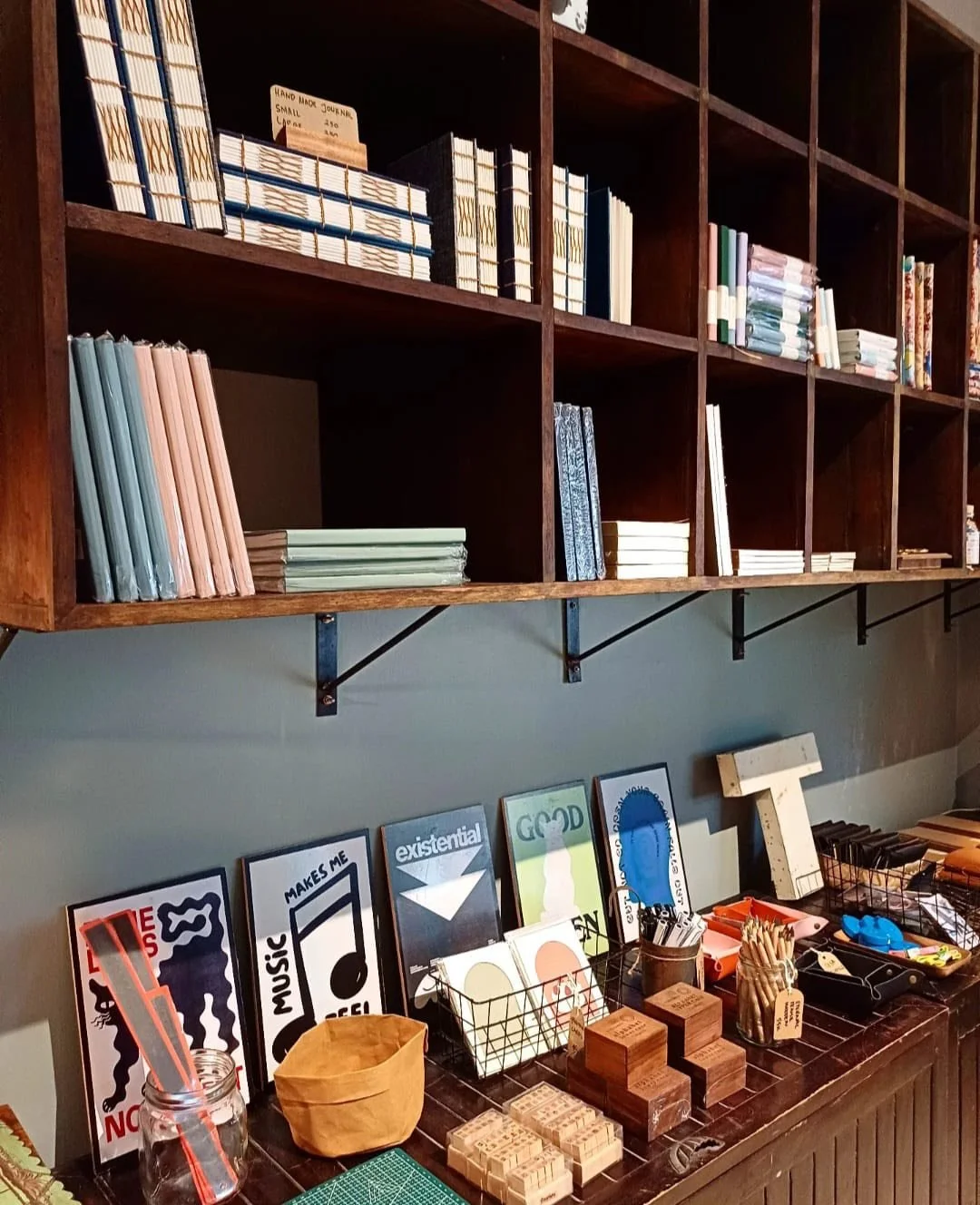 Wooden shelves filled with neatly stacked notebooks and journals, with colorful artwork and various stationery items on a dark wooden table below.