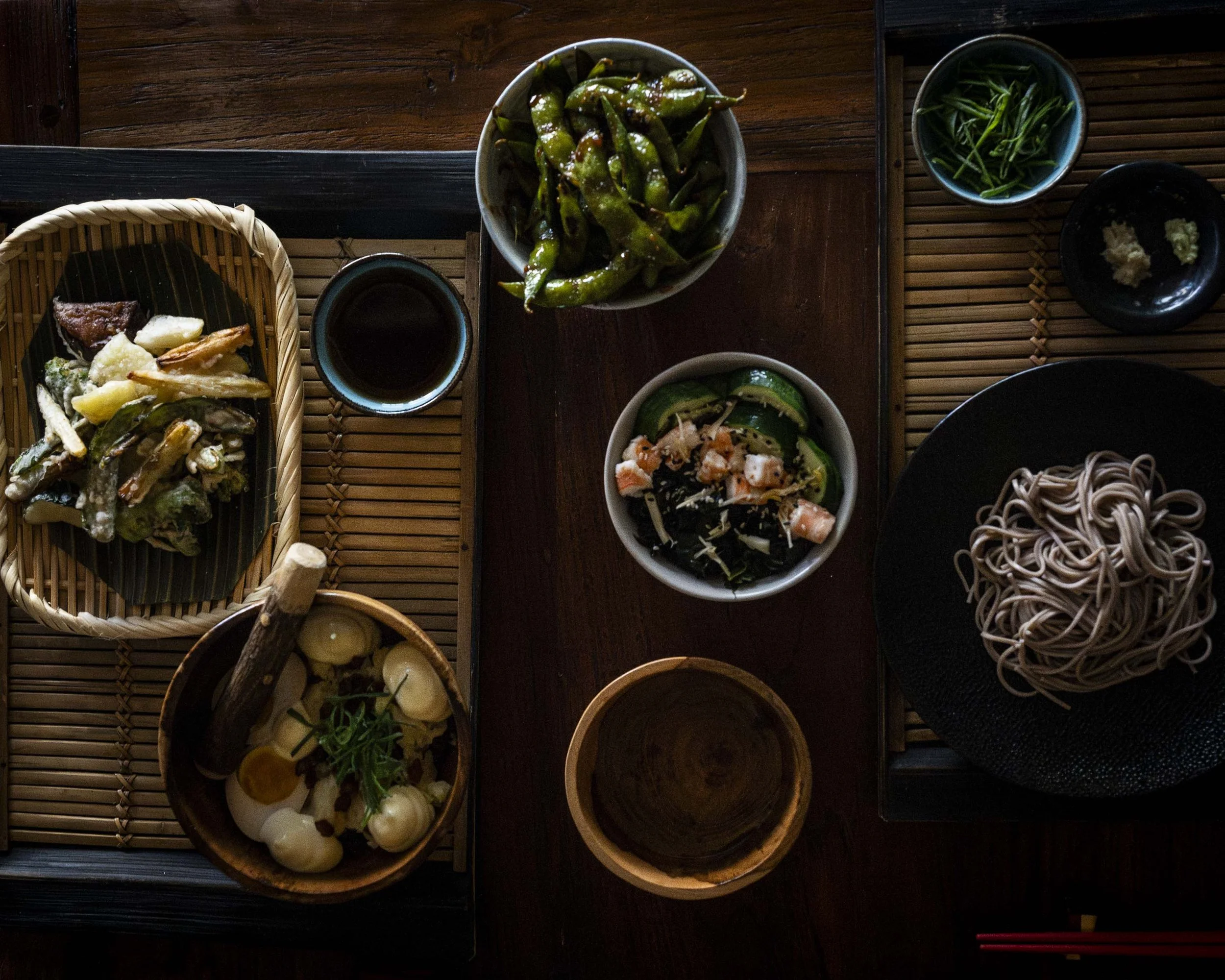 Japanese meal with soba noodles, edamame, tempura vegetables, pickled cucumbers, sliced rolled fish, and dipping sauce on a wooden table.