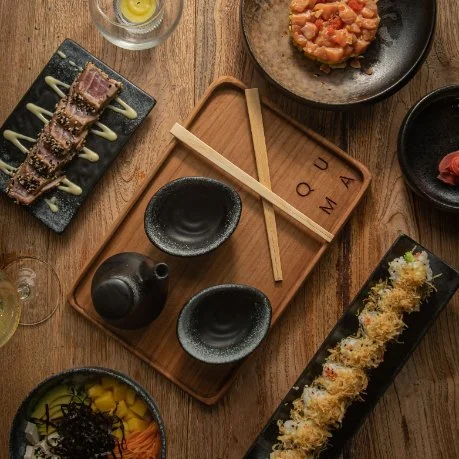 Various Japanese dishes on a wooden table, including sushi rolls, a bowl of rice with seaweed, a soy sauce container, and a drink.
