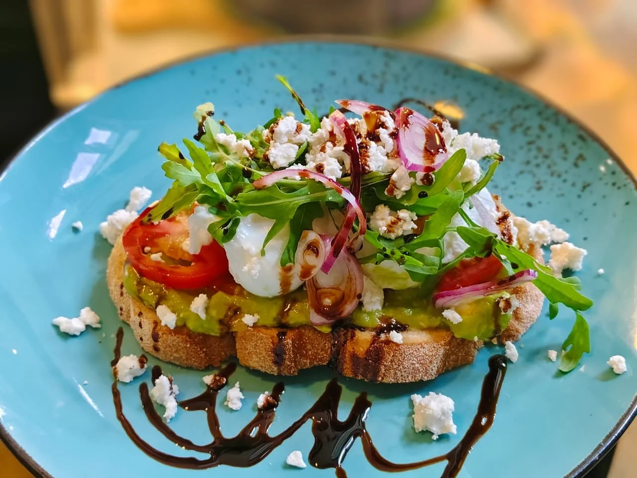 Open-faced toast with avocado, tomato, poached egg, arugula, red onion, crumbled cheese, balsamic glaze, and chocolate drizzle on a blue plate