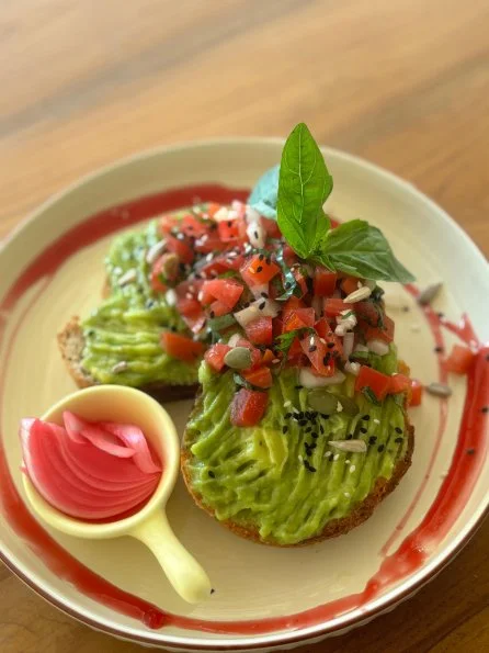 Two slices of avocado toast topped with tomato salsa and a basil leaf, served with pickled pink onions on a decorated plate.