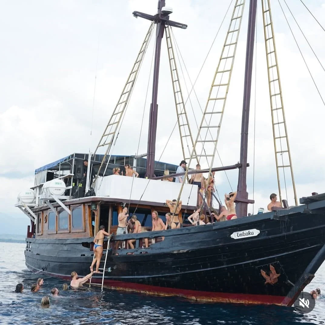 A group of people swimming and lounging on a boat named 'Emboku' on the water, with some on the deck and others in the water near the boat.