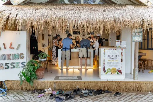 Group of people at an outdoor bar with a thatched roof, engaged in conversation, with shoes on the ground outside and signs indicating cooking classes.