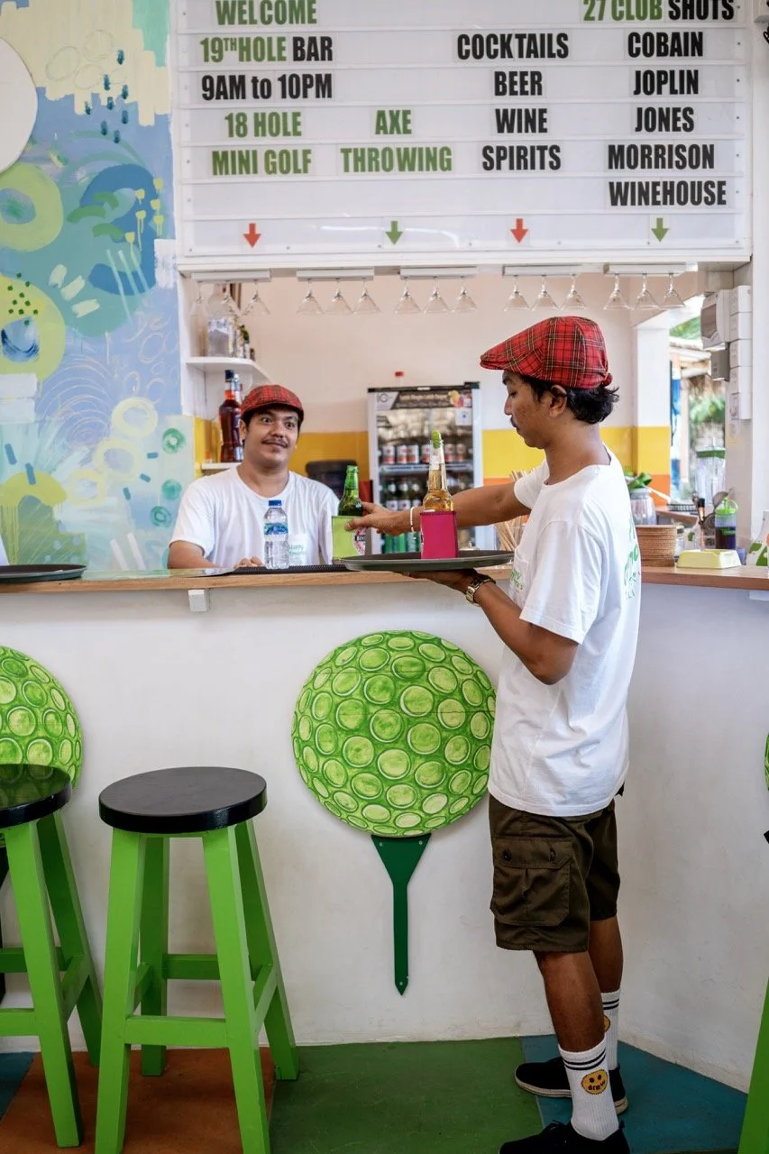 Two young men in white t-shirts and red plaid hats at a bar counter with bright green bar stools designed like golf balls. One is serving drinks while the other is smiling behind the counter. A colorful decorator wall and a menu board with drink options are visible in the background.