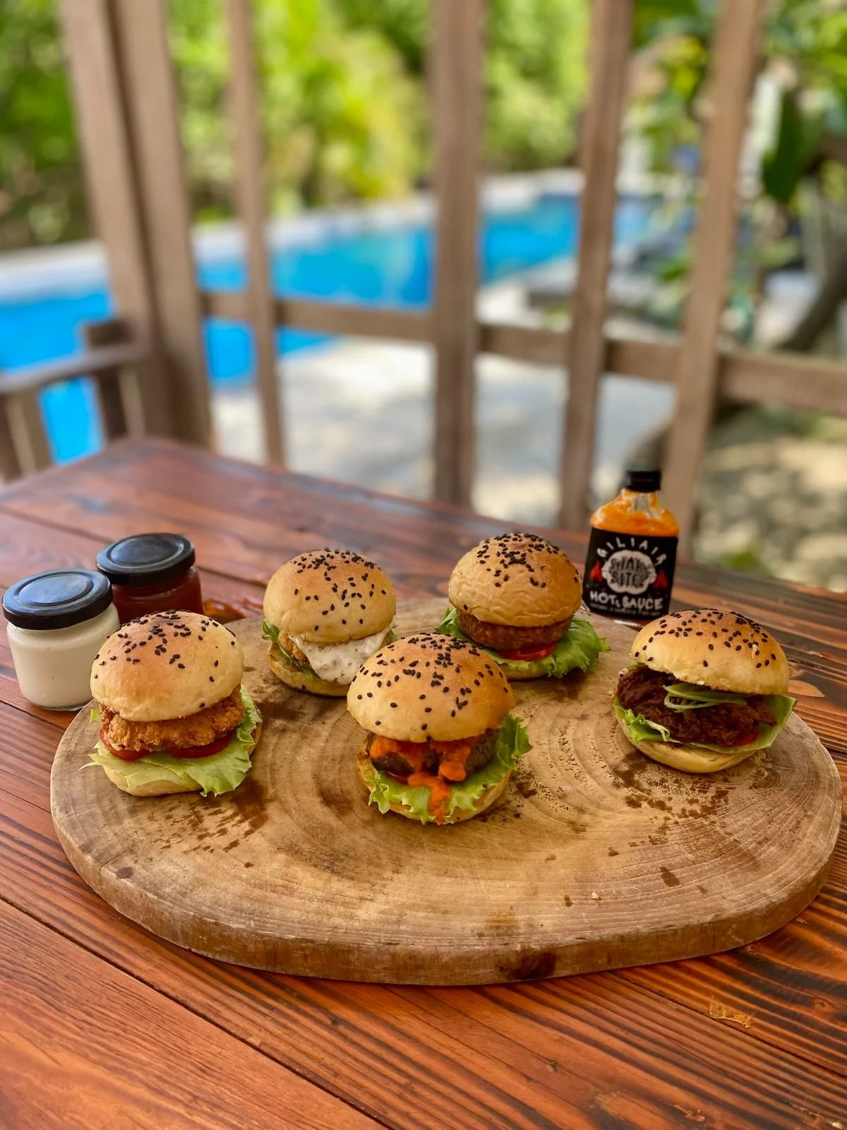 Five mini burgers with various toppings on a wooden round board, with mustard, ketchup, and mayonnaise in small jars, and a bottle of hot sauce, on a wooden table with a backyard in the background.