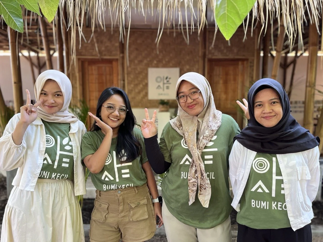 Four women wearing green T-shirts with white text and symbols, standing in front of a bamboo and thatched structure, smiling and posing for the photo. Two women are wearing hijabs, and one has glasses.
