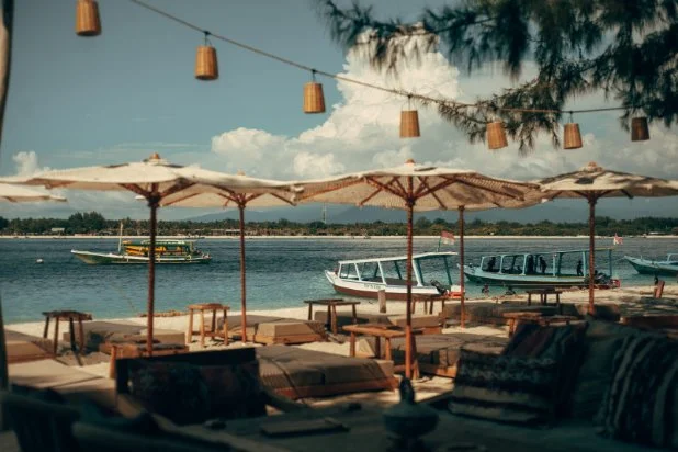Beachside seating area with umbrellas and tables overlooking a river, with boats passing by and a tree in the foreground.