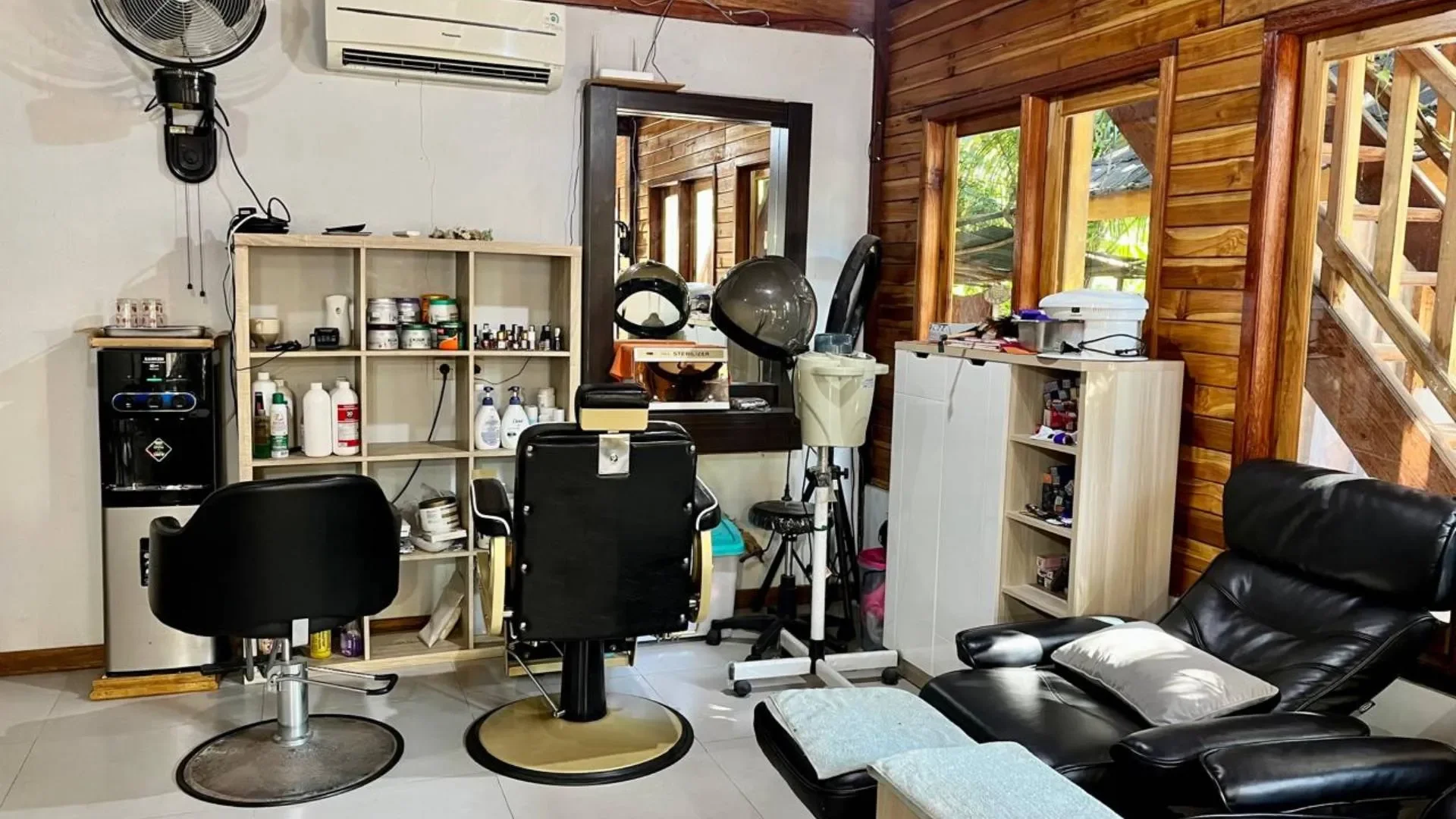 A salon or spa room with a black salon chair in front of a mirror, shelves with hair and nail products, a black leather recliner with a pillow, and large windows with wooden frames letting in natural light.