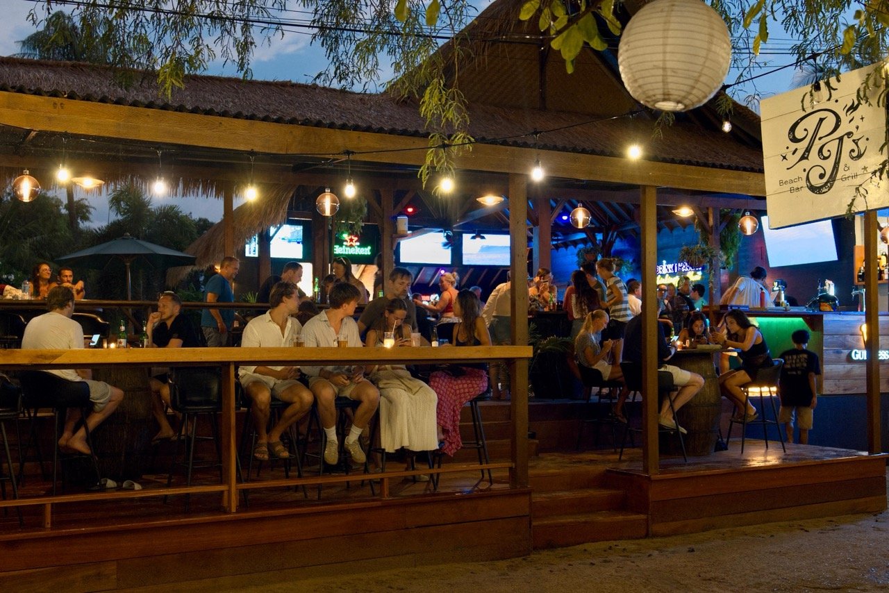 People socializing and drinking at an outdoor bar in the evening, with string lights, a thatched roof, and a bar counter illuminated with green lighting.