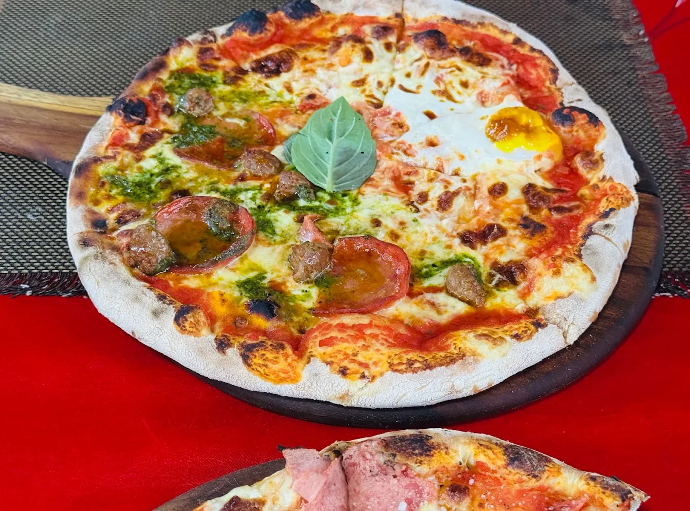 A pizza topped with sausage, pepperoni, basil leaves, and a sunny side up egg, served on a wooden pizza board with a red tablecloth.